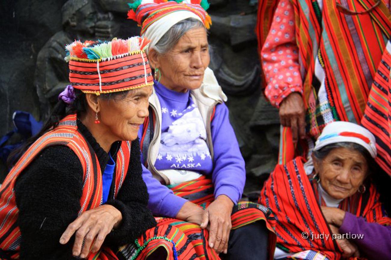 A group of Indigenous people in traditional clothing kneeling and sitting around each other